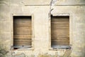 Two brown grungy yellow wooden windows in a gray yellow broken cracked facade of an abandoned derelict house