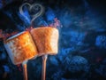 Two brown sweet marshmallows roasting over fire flames. Smoke in form of hearts. Marshmallow on skewers roasted on Royalty Free Stock Photo