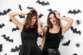 Two brunette girls dressed in black dresses grimace on the background of the wall with bats. Halloween party. Royalty Free Stock Photo