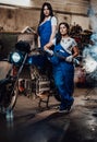 Two brunette women in blue overalls posing next to a custom bobber in authentic workshop garage Royalty Free Stock Photo