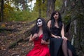 Two brunettes women with makeup like a Halloween skull and Halloween witch makeup sits in a red and black dresses on the roots