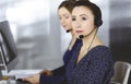 Two busineswomen have conversations with the clients by headsets, while sitting at the desk in a modern office. Diverse