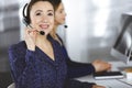 Two busineswomen have conversations with the clients by headsets, while sitting at the desk in a modern office. Diverse