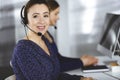 Two busineswomen have conversations with the clients by headsets, while sitting at the desk in a modern office. Diverse