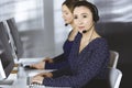 Two busineswomen are talking to the clients by headsets, while sitting at the desk in a modern office. Diverse people