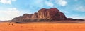 Two camels, walking on orange red sand of Wadi Rum desert, large mountains with blue sky above background