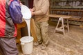 Two carpentry workers pour wood glue from a large plastic bucket into another empty