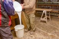 Two carpentry workers pour wood glue from a large plastic bucket into another empty