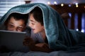 Two caucasian children holding digital tablet while lying under blanket in the dark at night reading online book