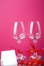 Two champagne glasses with ribbons on a pink background and and a card and a heart candlestick with a burning candle.