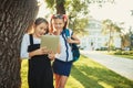 Two charming school-age girls stand by the tree near the school and play on the tablet