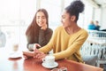 Two cheerful and beautiful girls are sitting together near the table and watching something on the phone. They look Royalty Free Stock Photo