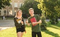 Two cheerful friends, students with broad smiles holding textbooks and coffee in the park near the high school building. Young