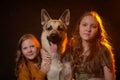 Two cheerful joyfull sisters having fun together and with big german shepherd dog in dark studio with black background