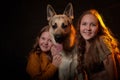 Two cheerful joyfull sisters having fun together and with big german shepherd dog in dark studio with black background