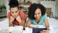 Two cheerful mixed race funny girlfriends reading book and using tablet computer talks and have fun lying in bed at home Royalty Free Stock Photo