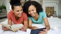 Two cheerful mixed race funny girlfriends reading book and using tablet computer talks and have fun lying in bed at home Royalty Free Stock Photo