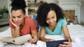 Two cheerful mixed race funny girlfriends reading book and using tablet computer talks and have fun lying in bed at home Royalty Free Stock Photo