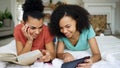 Two cheerful mixed race funny girlfriends reading book and using tablet computer talks and have fun lying in bed at home Royalty Free Stock Photo