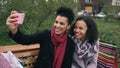 Two cheerful mixed race woman with shopping bags talking at video call with smartpone. Young girls sitting on bench have Royalty Free Stock Photo