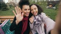 Two cheerful mixed race woman with shopping bags talking at video call with smartpone. Young girls sitting on bench have Royalty Free Stock Photo