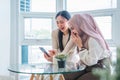 Two cheerful multi ethnic young women looking at phone together while sitting indoors in cafe, talking and laughing. Royalty Free Stock Photo