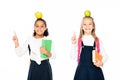 two cheerful multicultural schoolgirls with apples on heads showing idea gestures isolated on white. Royalty Free Stock Photo