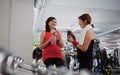 Two senior women in gym resting after doing exercise, holding water bottles.