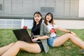 Two cheerful young asian girls friends looking at camera while sitting on the lawn outdoors with cups of coffee and Royalty Free Stock Photo