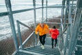 Two cheerfully smiling bright sporty clothes dressed men running up on huge steel industrial stairs with picturesque winter city