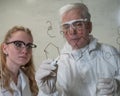 Two chemist colleagues write formulas on glass. Caucasian elderly man and young woman brainstorming. Royalty Free Stock Photo