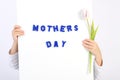 Two childhood hands holding white board with blue text Mothers day and one white and violet tulip Royalty Free Stock Photo