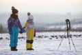 Two children boy and girl having fun outside in winter playing with photo camera on a tripod on snow covered field Royalty Free Stock Photo