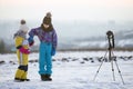 Two children boy and girl having fun outside in winter playing with photo camera on a tripod on snow covered field Royalty Free Stock Photo