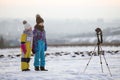 Two children boy and girl having fun outside in winter playing with photo camera on a tripod on snow covered field Royalty Free Stock Photo