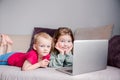 Two children a boy and a girl lie on a sofa in home clothes and watch videos on a laptop and look at camera for a moment Royalty Free Stock Photo