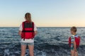 Two children standing in the beach wearing red life jackets at sunset. Sea gale and people in life vest on shore