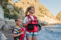 Two children in wearing red life jackets standing on the beach near the rocky seashore. Equipment for rescuing people and buoyancy