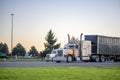 Two big rig classic semi trucks with turned on headlights with loaded semi trailers standing on the parking lot for rest take a Royalty Free Stock Photo