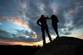 Silhouettes of man and woman look at sunset on the top of rock. Hikers couple on dramatic sky at sunset. Back view.