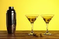 Two cocktails in martini glasses with green olives and shaker on a wooden surface against yellow background with copy space Royalty Free Stock Photo