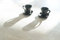 Two Coffee clay black cups on table with bright backlight which draws long cup shadows. Coffee for couple in modern kitchen