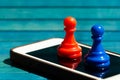 Two colorful chess pawns, red and blue colored game pieces standing on top of a modern smartphone screen, display closeup, mobile