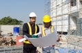 Two construction worker looking at a floor plan, standing in front of a large building site with helmet and safety gear. Day time Royalty Free Stock Photo