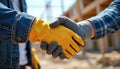 Two construction workers shake hands outdoors on a building site. They wear work gloves and protective gear. The image shows Royalty Free Stock Photo