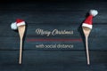Two cooking spoons with red Santa Claus hats, between them text Merry Christmas with social distance, dark blue wooden background Royalty Free Stock Photo