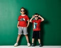 Two cool kids boys in modern summer wear shorts, red t-shirt with prints and sunglasses standing at green wall