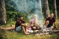 Two couples camping in woods. Men cooking sausages on campfire. Tired brunette lying on blond girls lap