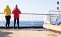 Two cruise passengers stand at the railing at the stern of a cruise ship and look back at the open sea