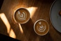 Two cups of fresh cappuccino with lovely latte art on brown wooden table with shadows and sun spots. Morning coffee for